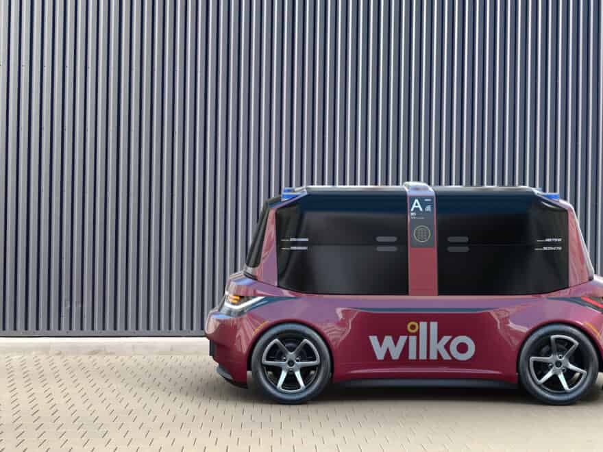 Side view of Pix-E small road-going electric delivery vehicle, pictured Rear view of Pix-E small road-going electric delivery vehicle, pictured in burgundy livery of omnichannel retailer Wilko, against vertical-striped grey fence.