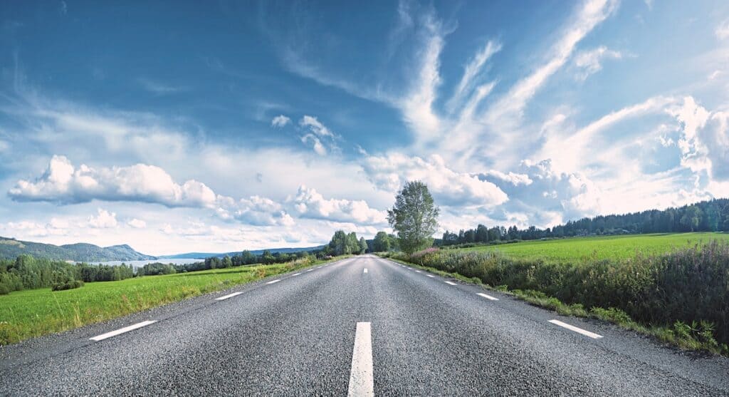 View down a straight empty road, with central white line markings, running into the horizon between fields in Sweden.