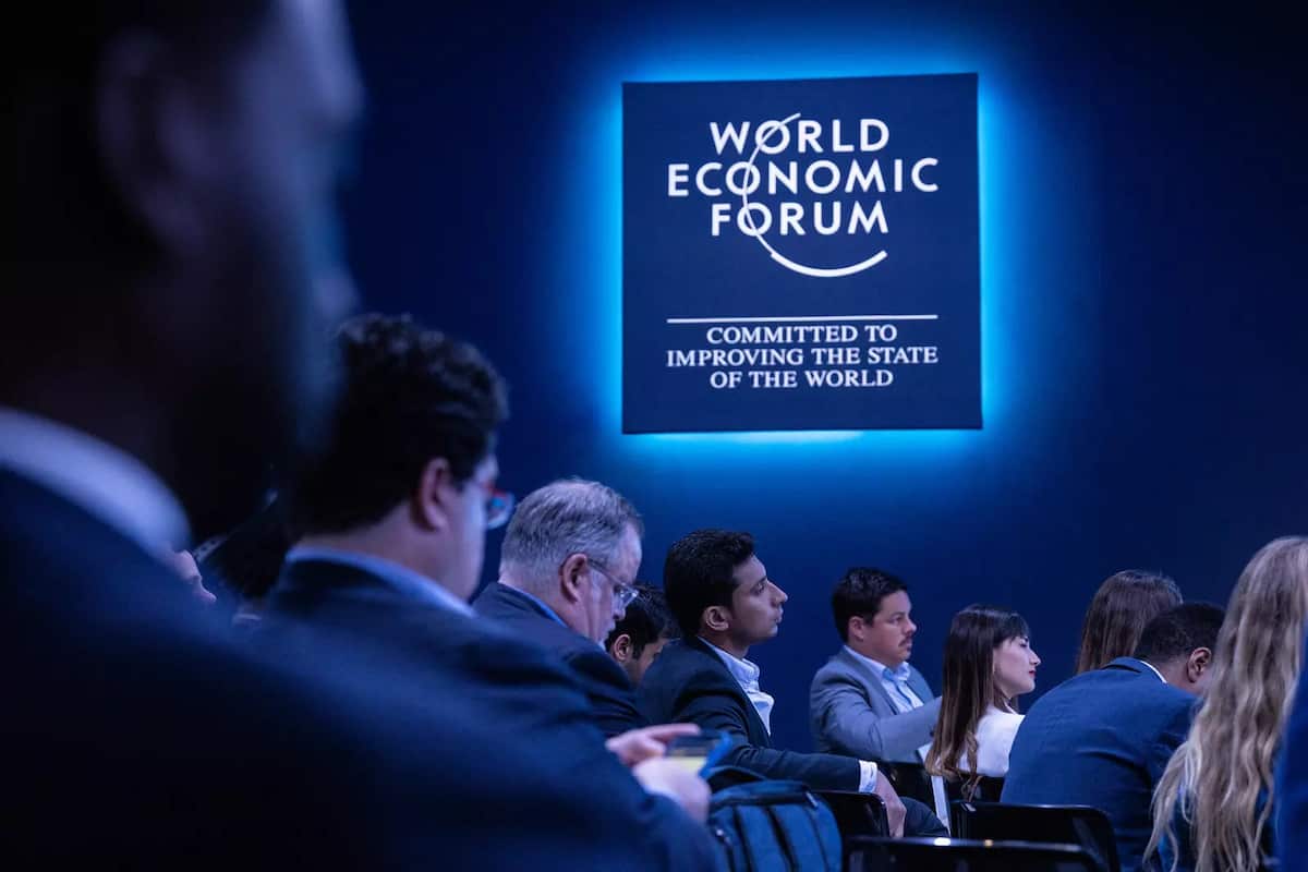 Business people seated in conference beneath illuminated sign: "World Economic Forum Committed to improving the state of the world." (Blue filter on image)