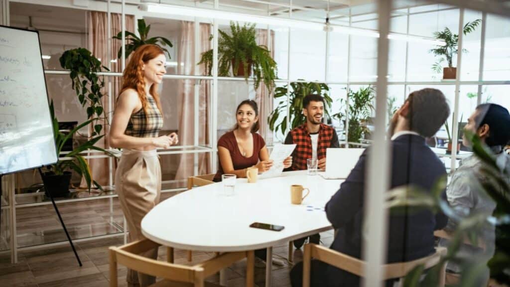 Image of diverse group of young professionals sat around an office table with one colleague stood presenting beside a whiteboard.