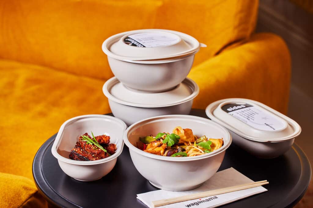 Tray with five new recyclable delivery bowls: three are round, two with lids stacked on top of one another, the third is open showing food; one oval bowl has lid, other displays food inside. Chopsticks and Wagamama branded napkins in foreground