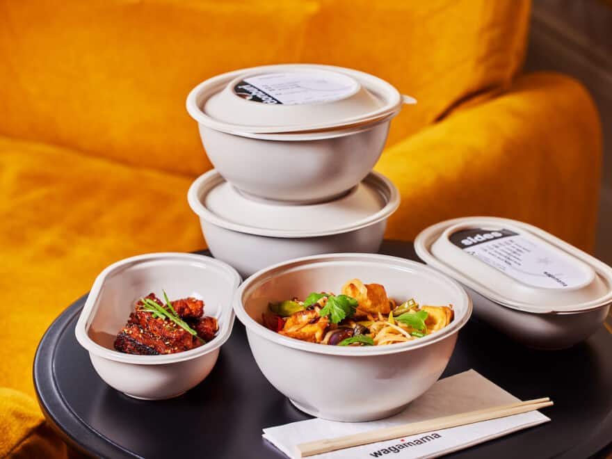 Tray with five new recyclable delivery bowls: three are round, two with lids stacked on top of one another, the third is open showing food; one oval bowl has lid, other displays food inside. Chopsticks and Wagamama branded napkins in foreground