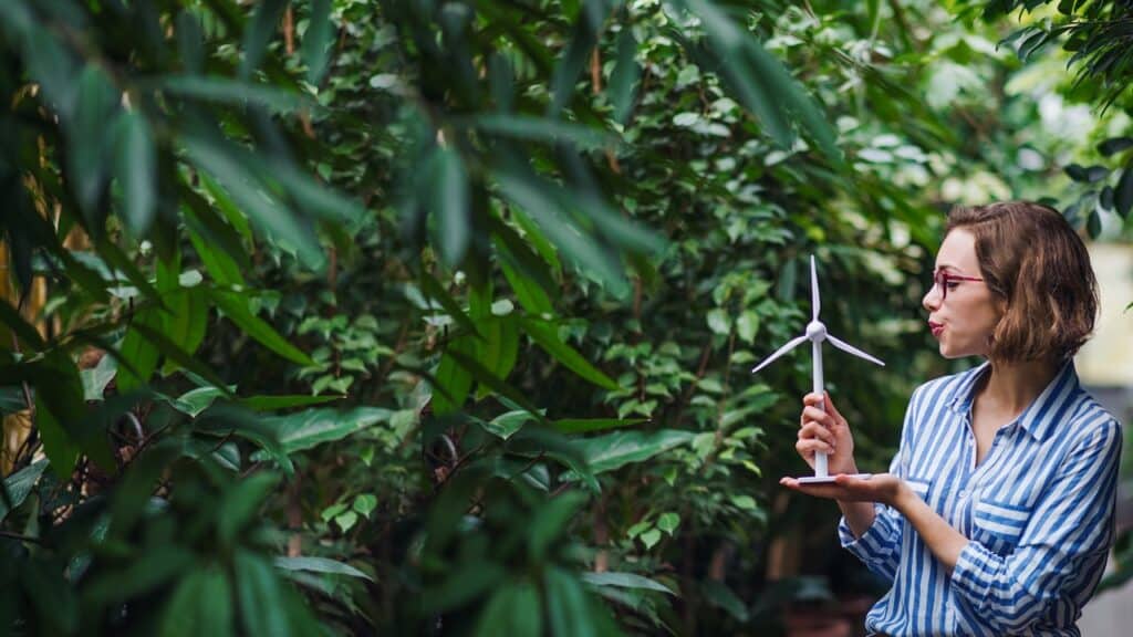 Person (female) pictured on right against backdrop of dense green foliage, blowing over model wind turbine they are holding.