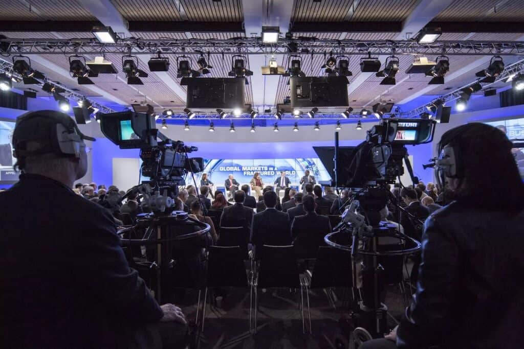 View from behind TV cameras at back of room towards stage at Davos conference panel session.