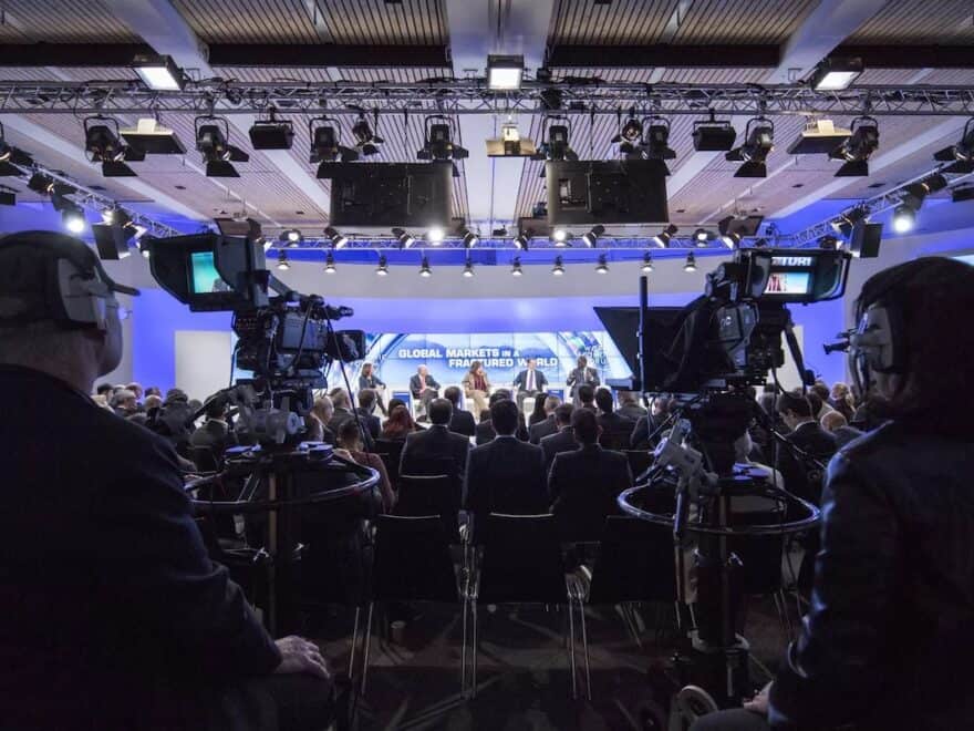 View from behind TV cameras at back of room towards stage at Davos conference panel session.