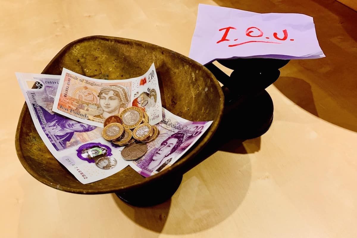 Weighing scales with notes and coins on one side and on the other a crumpled piece of white paper with I.O.U. written in red.