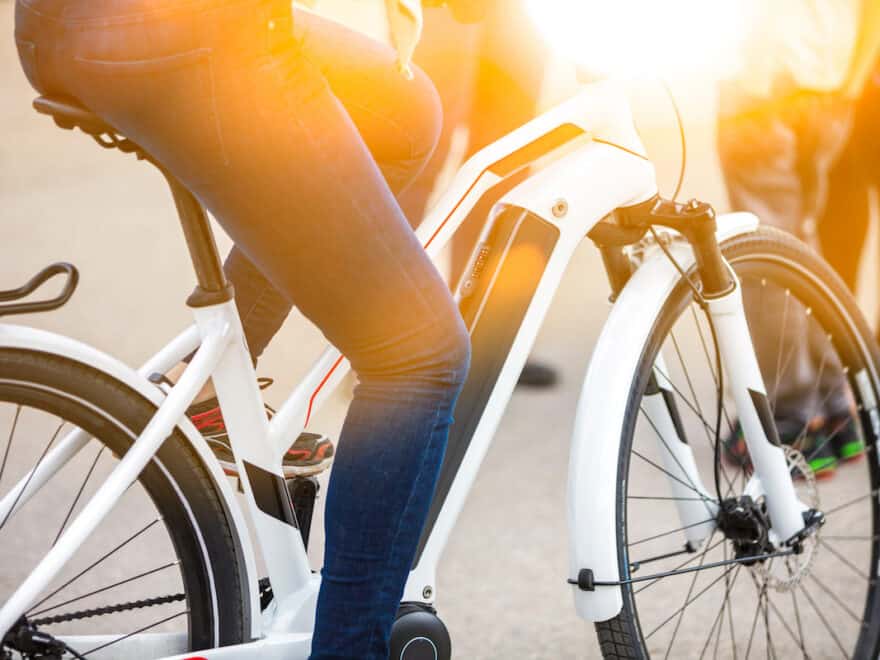 Close up of battery pack of a modern, white electric bicycle, being ridden by person wearing blue jeans, with legs only visible.