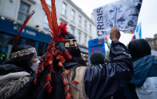 Indigenous peoples in crowd of protestors, wearing traditional headdress and photographed from behind, with banner above showing word 'CRISIS'.