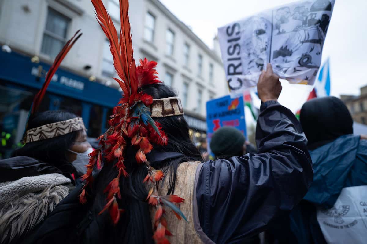 Indigenous peoples in crowd of protestors, wearing traditional headdress and photographed from behind, with banner above showing word 'CRISIS'.