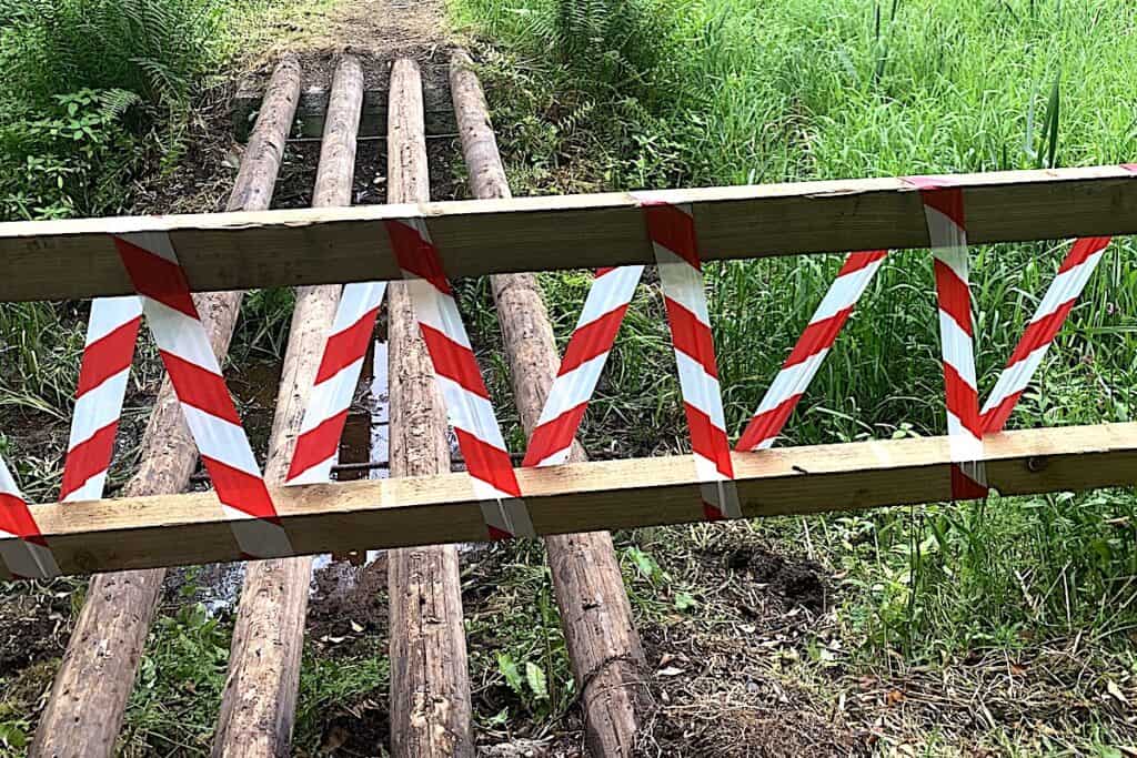 Red and white striped tape barrier across damaged wooden field bridge.