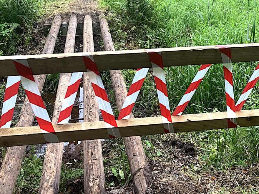 Red and white striped tape barrier across damaged wooden field bridge.