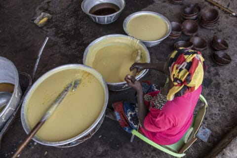 Woman viewed from above, seated in bright pink top, stirs one of three large pans of pale yellow custard-like shea.