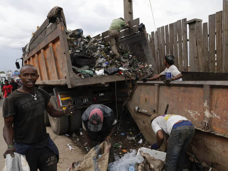 Men pictured picking up and sorting garbage from the street, beside rusty rubbish skip and lorry full of trash.