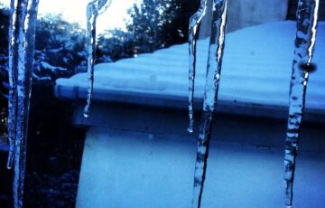 Icicles hanging down blue-filtered view out over snow-covered roof.