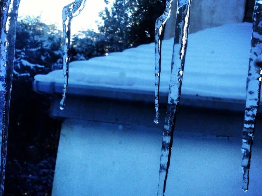 Icicles hanging down blue-filtered view out over snow-covered roof.
