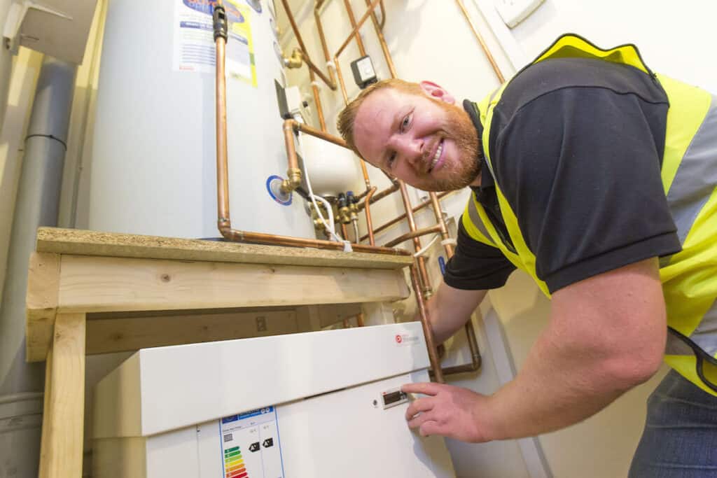 Smiling installation operative in yellow high-vis jacket presses button on Kensa Shoebox heat pump unit, pictured indoors with copper pipes above.