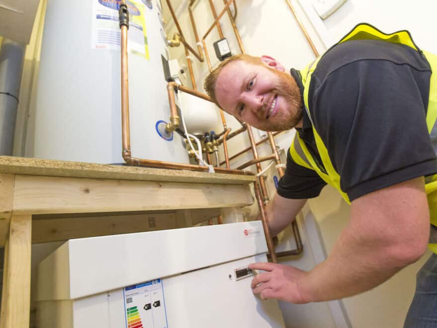 Smiling installation operative in yellow high-vis jacket presses button on Kensa Shoebox heat pump unit, pictured indoors with copper pipes above.
