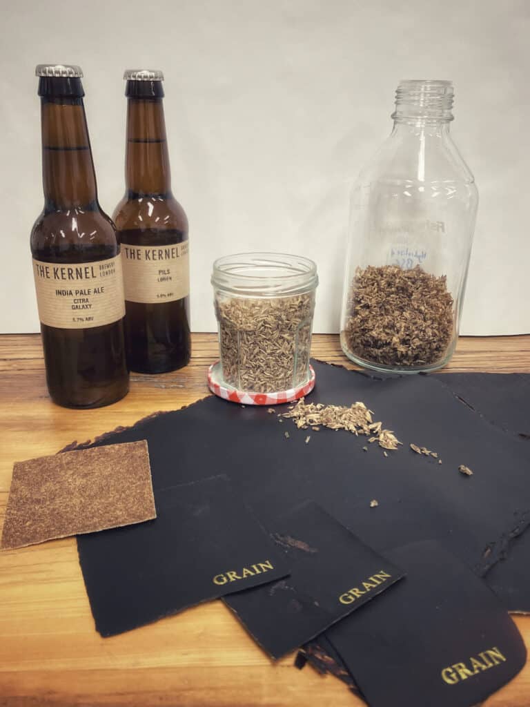 Brown bottles of The Kernel beer, black samples of leather alternative, with GRAIN branding in yellow, plus jar and bottle of spent and waste barley.