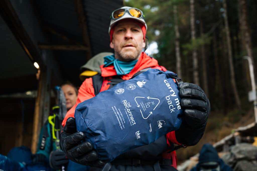 Person in climbing gear holding blue 'Carry me back' bag in front of them.