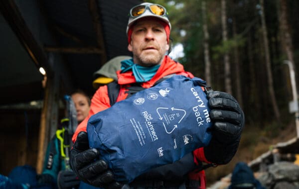 Person in climbing gear holding blue 'Carry me back' bag in front of them.