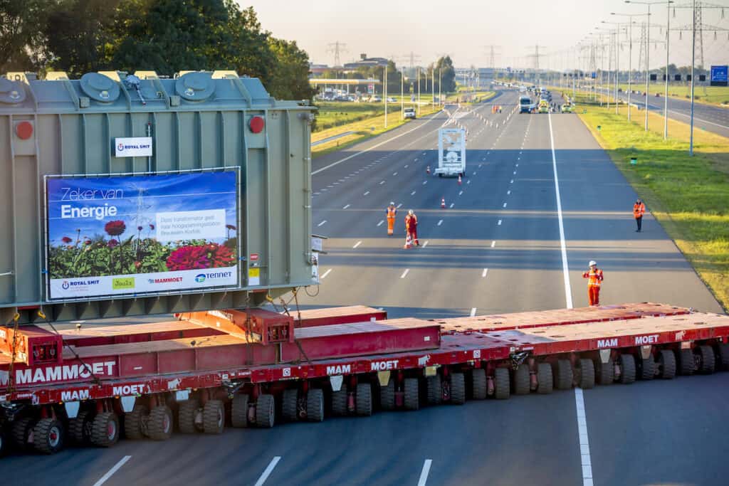 Multiple red modular transporters straddle closed-off highway, with Mammoet branding, carrying grey 'Zeker van Energie' transformer unit.