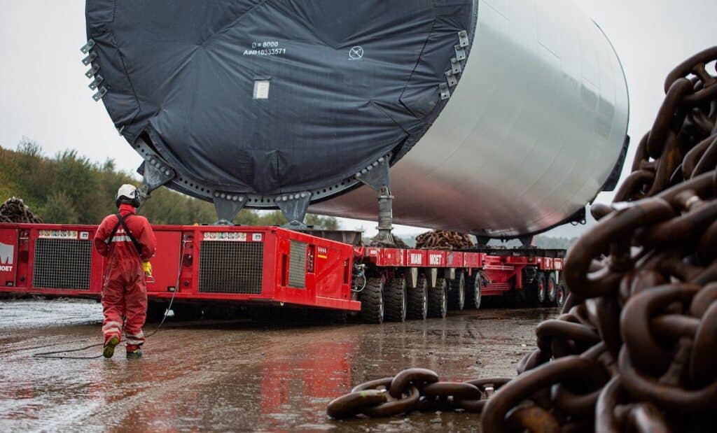 Red wheeled modular transporters carrying enormous cylindrical steel component, viewed end-on, with heavy metal chains in foreground.