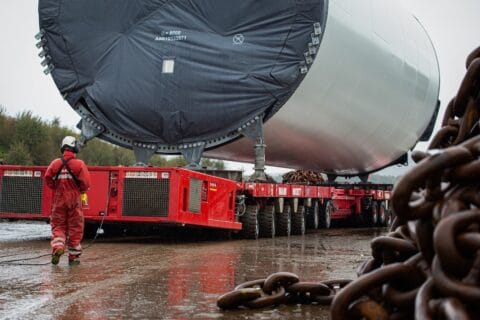 Red wheeled modular transporters carrying enormous cylindrical steel component, viewed end-on, with heavy metal chains in foreground.