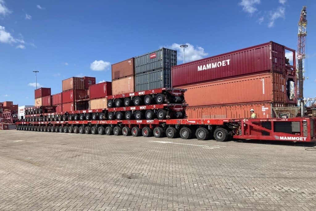 Multiple red modular transporters seen from side, piled high with 17 shipping containers, Mammoet branding visible, in concrete block yard.