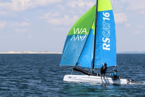 Racing catamaran on open sea with two crew, plus blue and green sails marked RSCAT16.