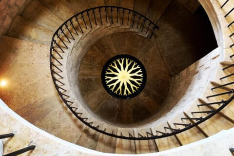 View down large stone spiral staircase, with bright yellow circular wheel design in centre at at bottom.