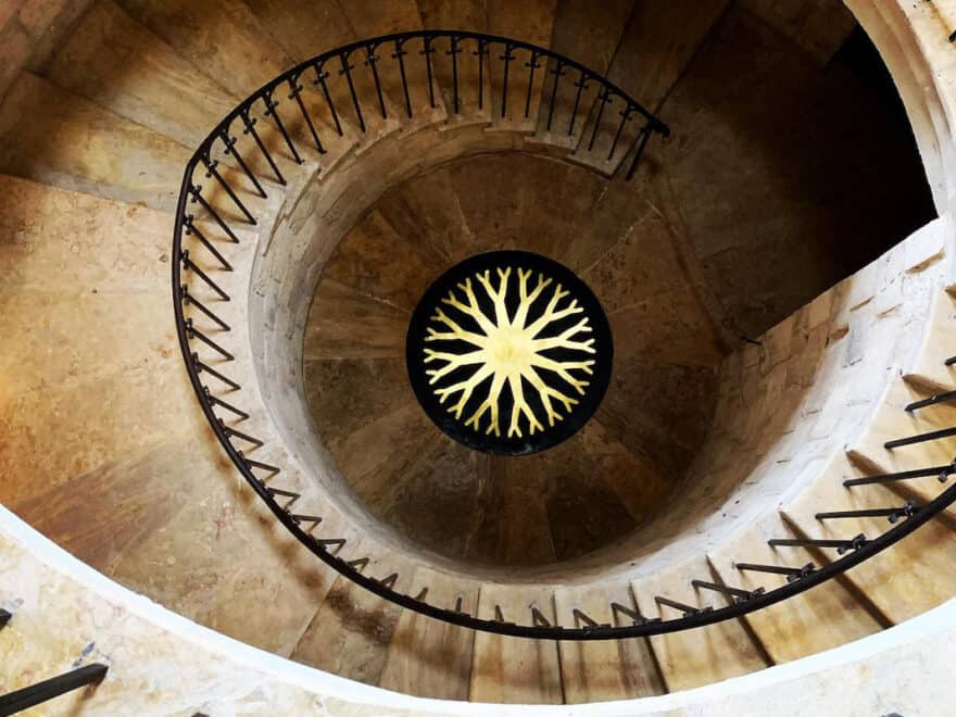 View down large stone spiral staircase, with bright yellow circular wheel design in centre at at bottom.