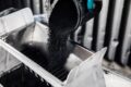 Mostly monochrome image of carbon black powder being poured out of a beaker in a laboratory, with thumb tip of cyan blue rubber glove visible.