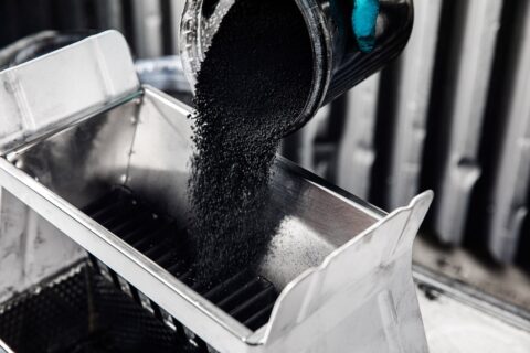 Mostly monochrome image of carbon black powder being poured out of a beaker in a laboratory, with thumb tip of cyan blue rubber glove visible.