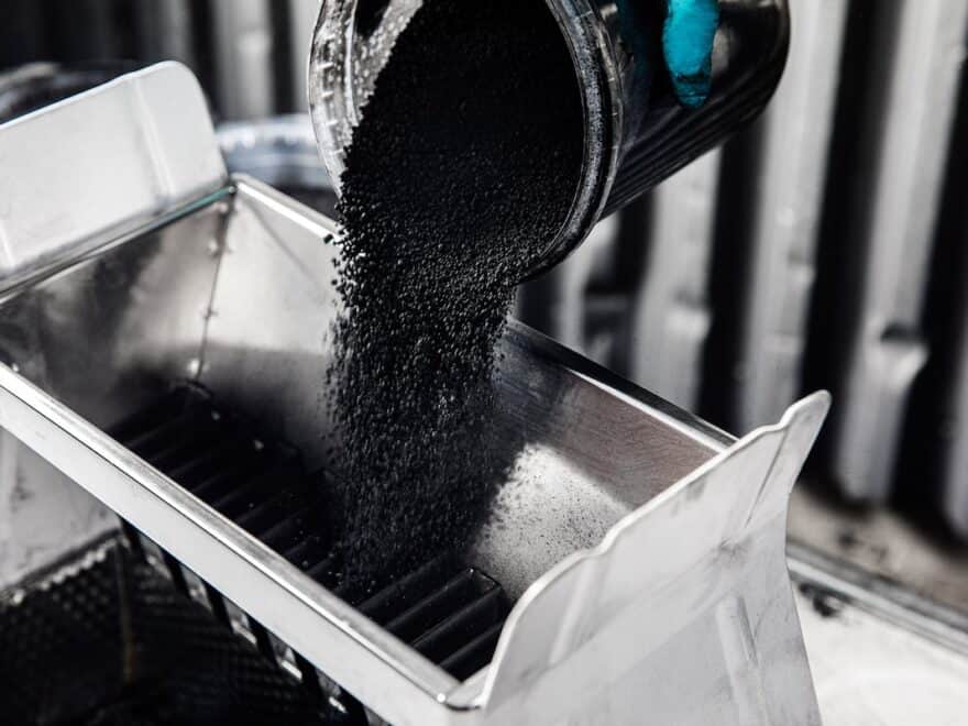 Mostly monochrome image of carbon black powder being poured out of a beaker in a laboratory, with thumb tip of cyan blue rubber glove visible.