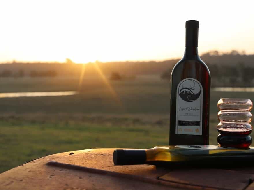 Two wine bottles, one upright, one lying flat, pictured with a glass of red wine against backdrop of sun setting over green fields.
