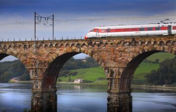 LNER train in red and white livery, pictured crossing stone bridge over water in countryside near Berwick.