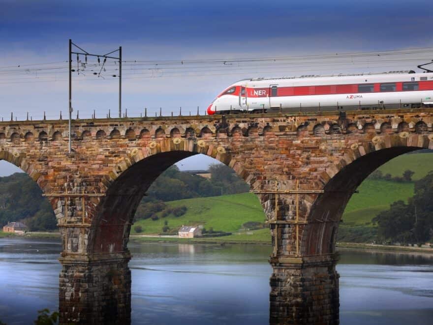 LNER train in red and white livery, pictured crossing stone bridge over water in countryside near Berwick.