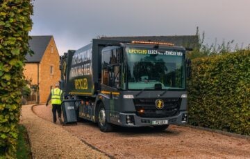 Upcycled electric vehicle (UEV) refuse truck in grey, pictured on street of quiet new residential development, with green hedges either side.
