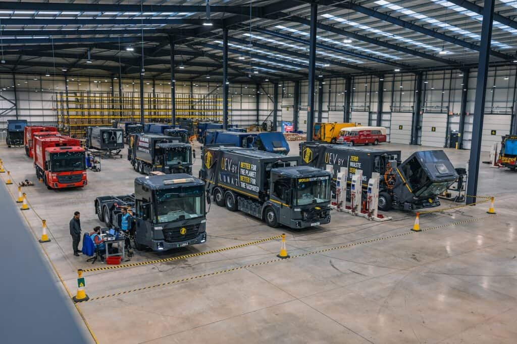 Numerous refuse trucks, most in grey livery, some red, pictured in process of being upcycled, parked on workshop floor in large hangar.