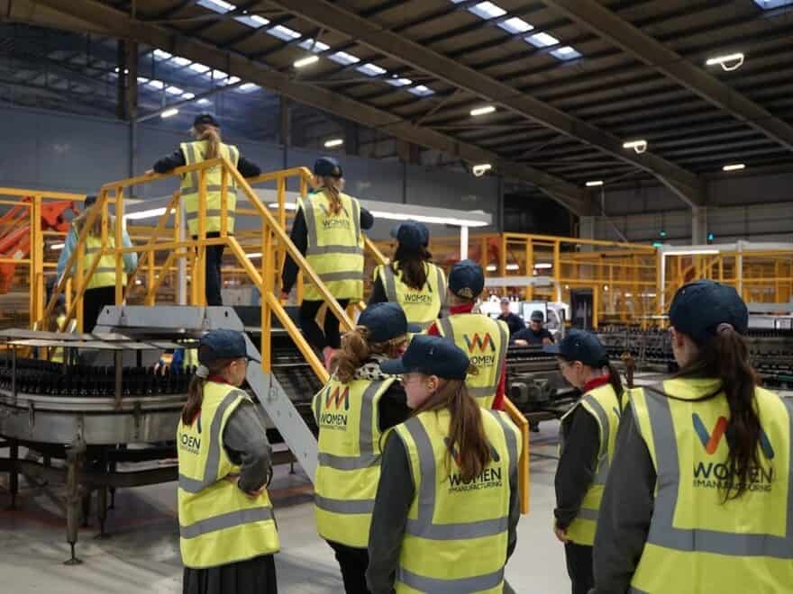 Women seen climbing metal stair in factory setting, each wearing a yellow high-vis jacket, with 'Women in Manufacturing' written on back.