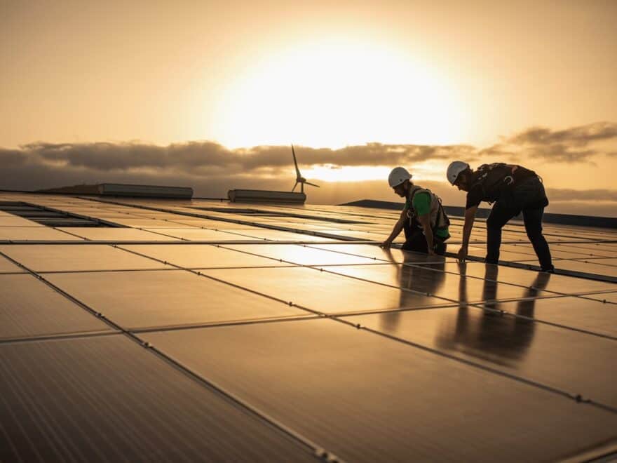 Workers in safety gear and hard hats on solar panel roof at sunset, with wind turbine in background.