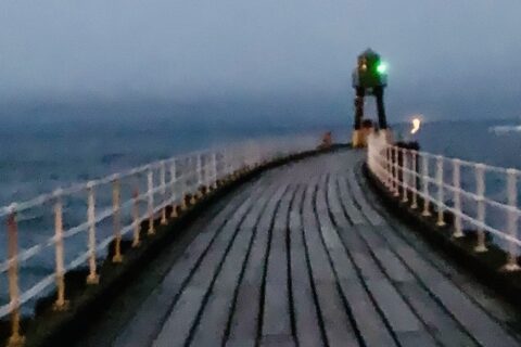 Diminishing artsy perspective, looking along the length of a wooden pier, towards a green light, with a red emergency lifebelt in the foreground.