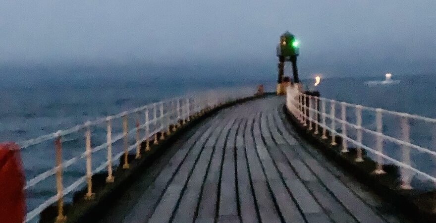 Diminishing artsy perspective, looking along the length of a wooden pier, towards a green light, with a red emergency lifebelt in the foreground.