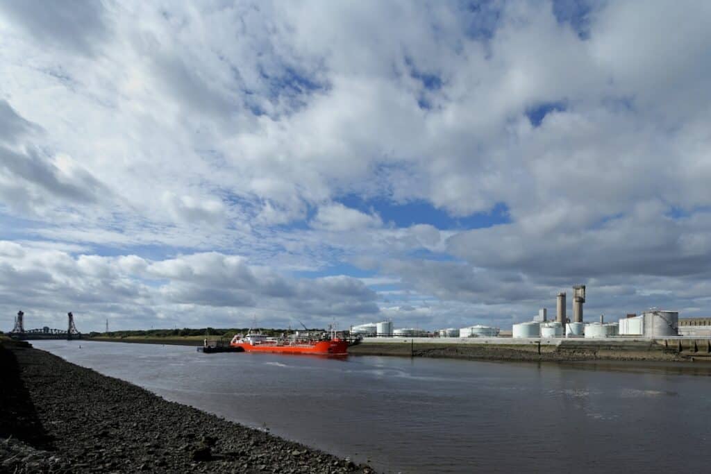 View of Riverside site from across the water, showing industrial facilities and planned development area, with bright oranger cargo ship alongside.