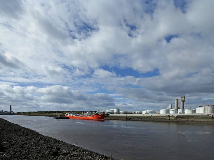 View of Riverside site from across the water, showing industrial facilities and planned development area, with bright oranger cargo ship alongside.