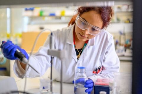 Portrait of PhD researcher Susana Meza Huaman working in the lab, in white coat, with protective glasses.