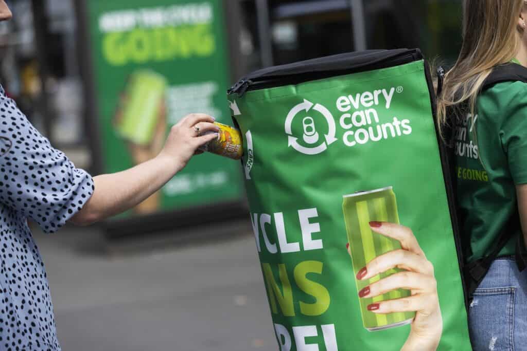 Person approaching from behind to put can into branded 'Every can counts' recycling backpack, being worn by another.