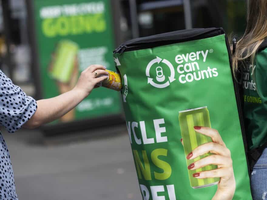 Person approaching from behind to put can into branded 'Every can counts' recycling backpack, being worn by another.