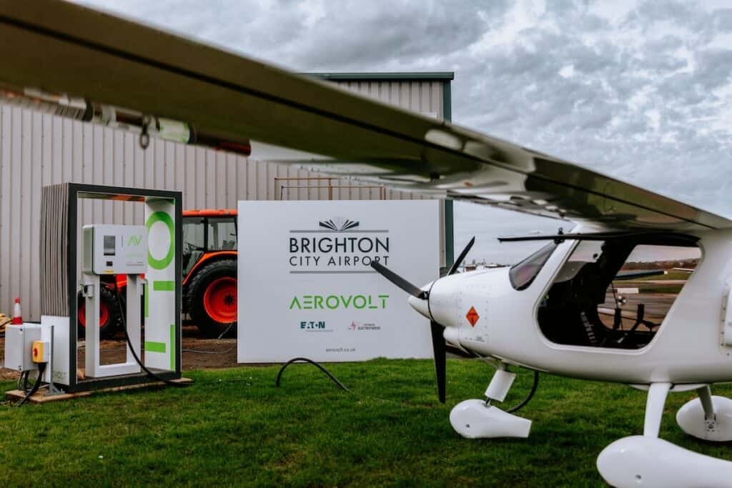 Small white 2-seater electric light aircraft pictured connected to airside charging station, beside sign for Brighton City Airport AeroVolt.