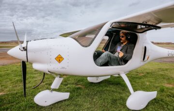 Small white 2-seater electric light aircraft pictured on grass, with cockpit door open, showing pilot holding Octopus Electroverse charging card.